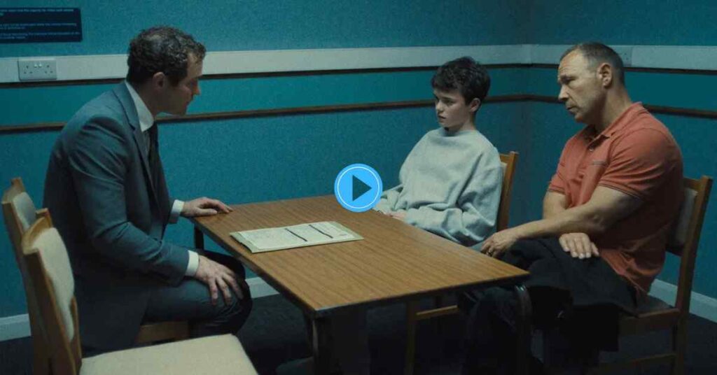 A man in a suit speaking to a young boy and an older man during a serious meeting, possibly an interrogation, in a TV drama.