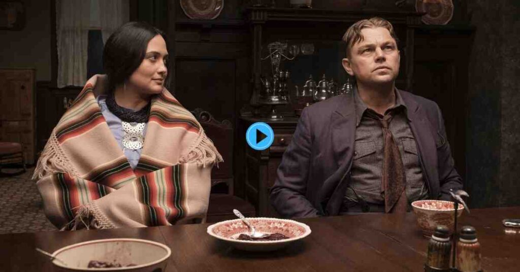 The two main actors from Killers of the Flower Moon sitting at a dark wood table with antique dishes in a dimly lit, wood-paneled room, with the woman wearing traditional Osage attire.