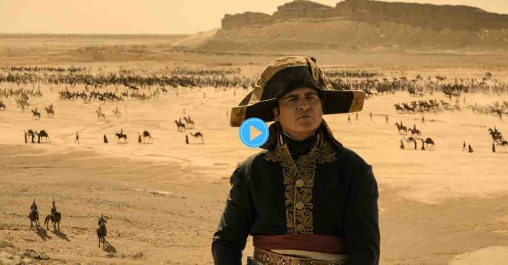 Joaquin Phoenix as Napoleon Bonaparte in his signature bicorne hat and uniform, standing on a dune overlooking a massive battle involving cavalry and camels in the desert.