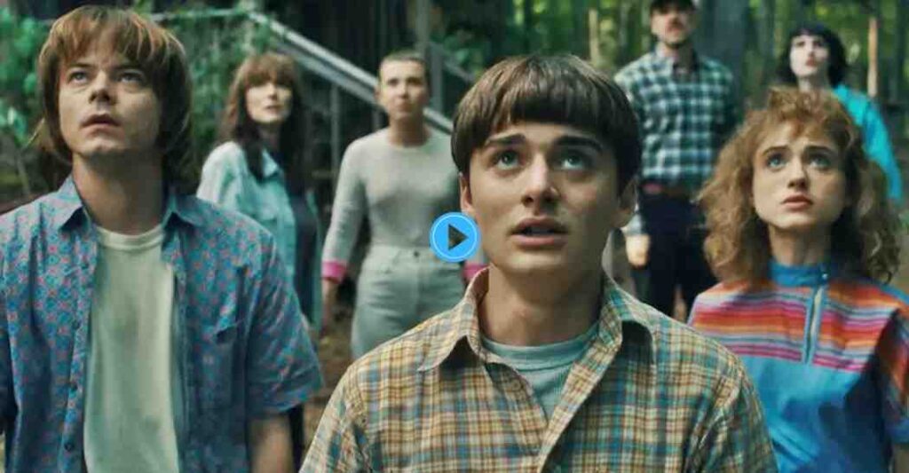 A large group of the main characters from Stranger Things stand in a forest looking up with concerned expressions.