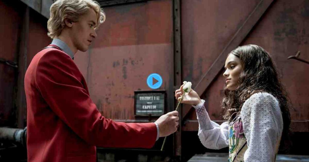 A still from The Hunger Games: The Ballad of Songbirds & Snakes showing Tom Blyth as Coriolanus Snow in a red suit offering a white rose to Rachel Zegler as Lucy Gray Baird.