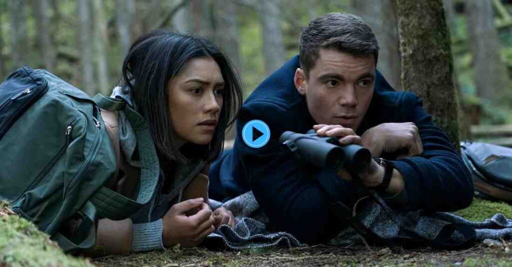 Gabriel Basso as Peter Sutherland and Luciane Buchanan as Rose Larkin lying low in a forest, with Peter looking through a pair of binoculars, in a scene from the series The Night Agent.