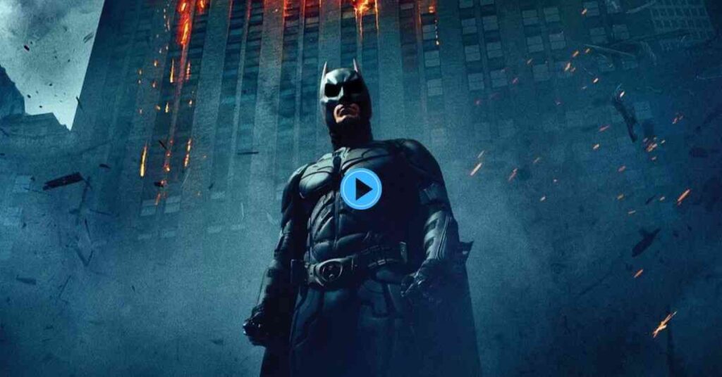 A dramatic low-angle shot of Batman (Christian Bale) standing against the backdrop of a massive, dark skyscraper with flames and sparks visible at the top, emphasizing a smoky, destroyed environment.